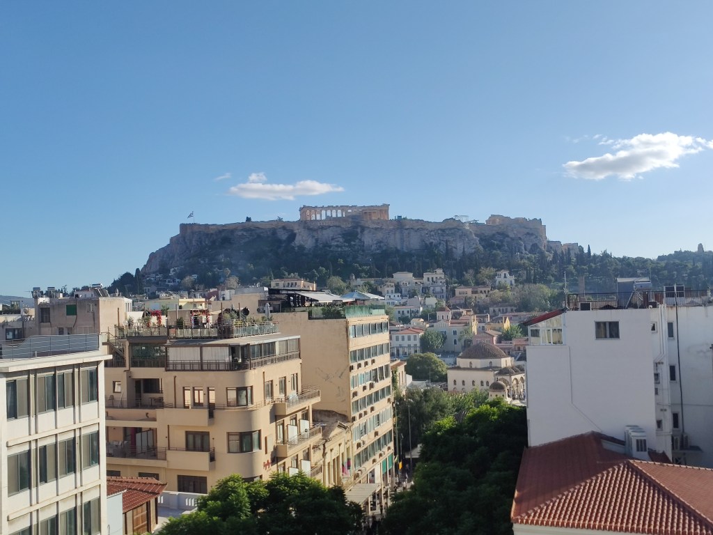 A rooftop balcony view in Athens overlooking city buildings and the Acropolis hill with the Parthenon visible under a clear blue sky.