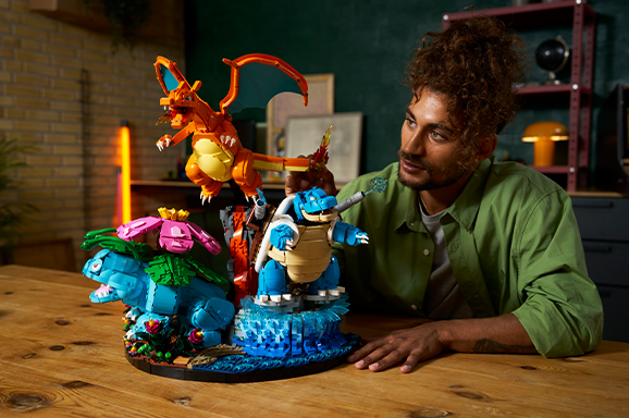 A LEGO Pokémon display featuring Charizard, Blastoise, and Venusaur being examined by a LEGO fan, showcasing the new LEGO Pokémon sets released in 2026.